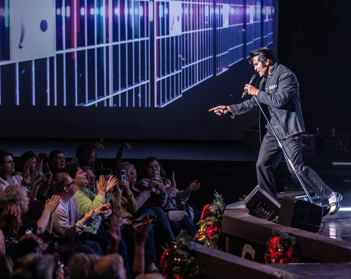An Elvis impersonator in a dark suit sings and points to an audience on stage with a digital prison cell background.
