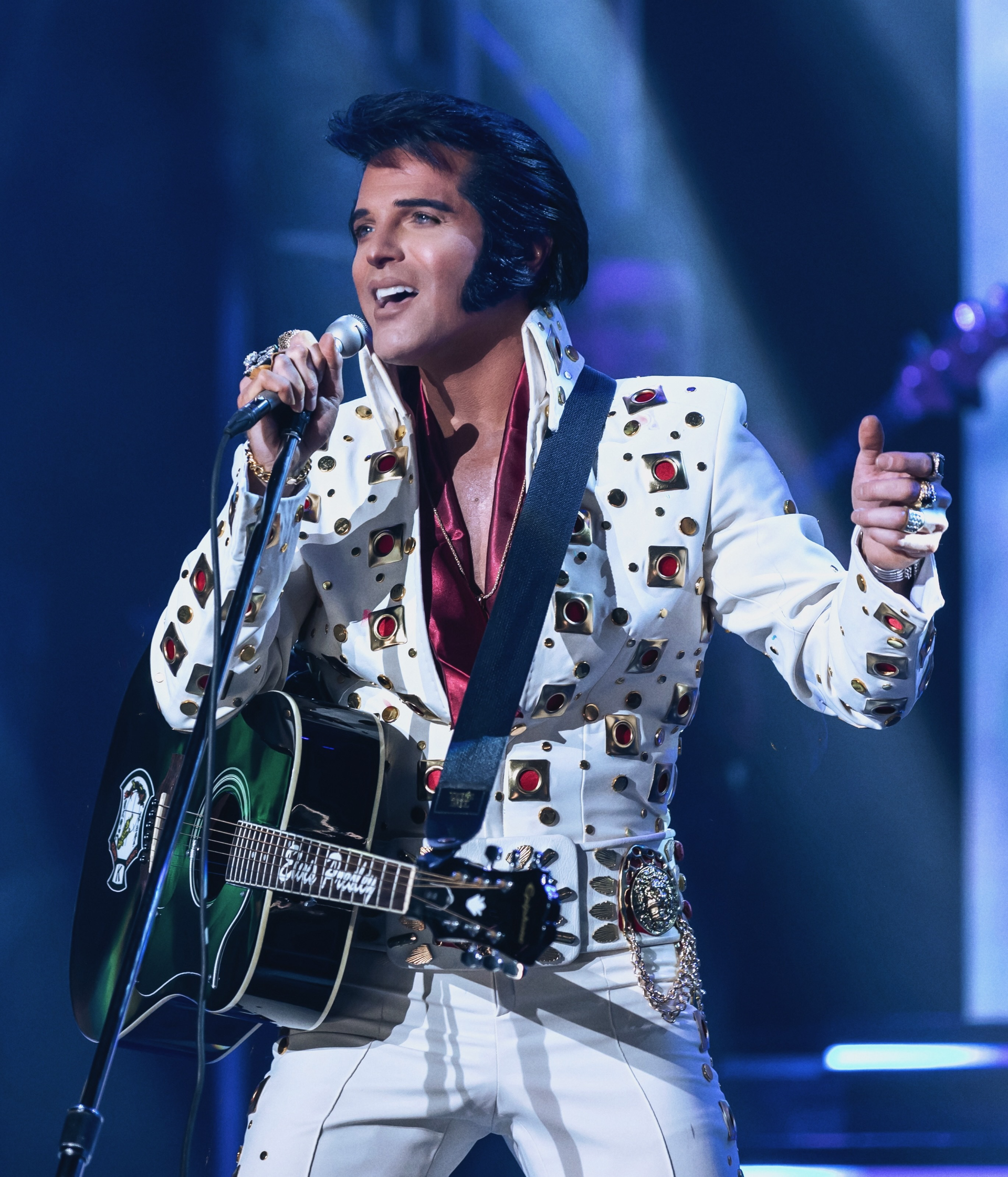 An Elvis Presley impersonator in a white, jewel-studded jumpsuit and cape, holding a guitar and microphone on stage.
