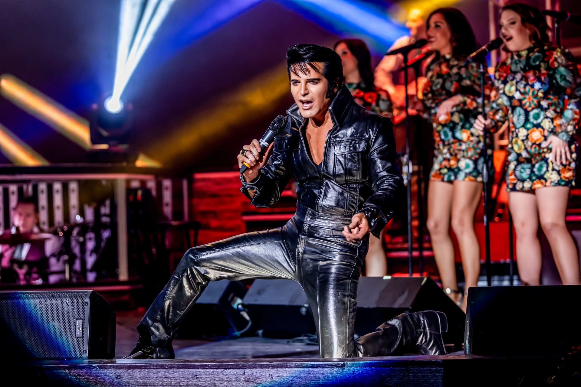 A performer in a black leather Elvis-style outfit sings into a microphone while kneeling on a stage with backup singers.