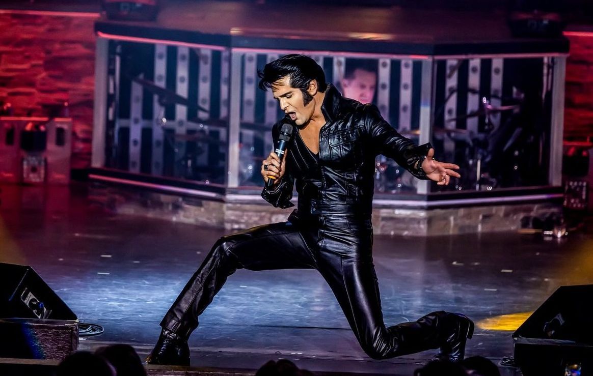 A performer dressed in a black leather outfit kneels on stage, singing into a microphone under spotlight.