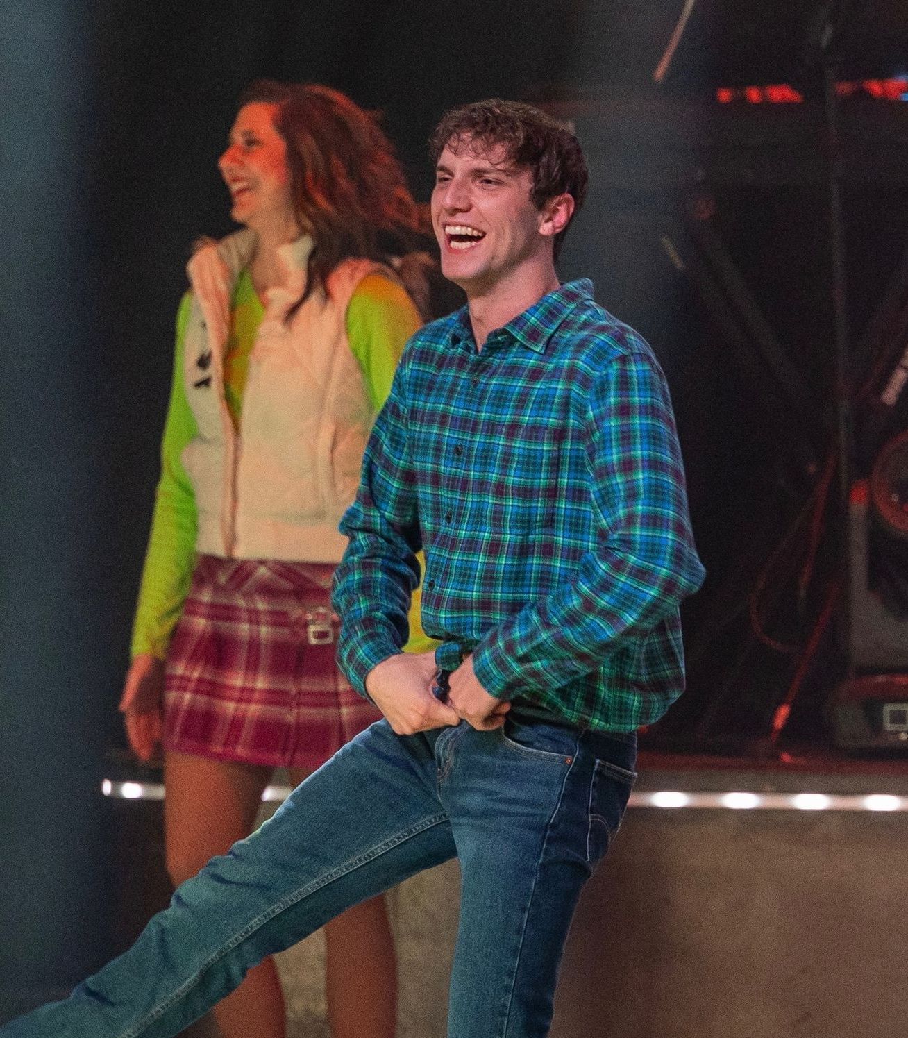 A man in a blue plaid shirt and jeans laughs while dancing on stage next to a woman in a green top, vest, and plaid skirt.