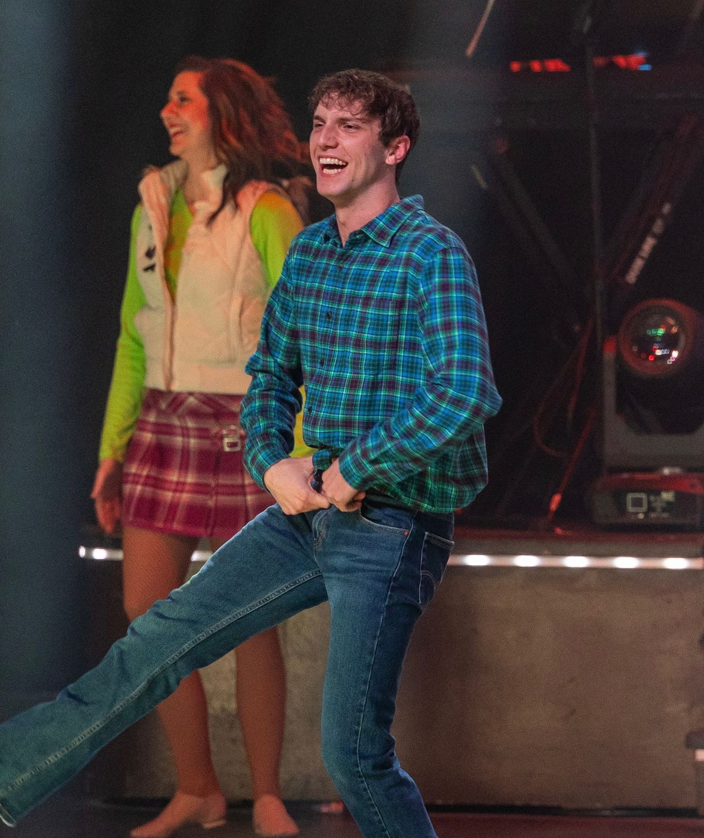 A smiling person in a blue plaid shirt and jeans kicks a leg, standing on a stage with a person in a vest and skirt.