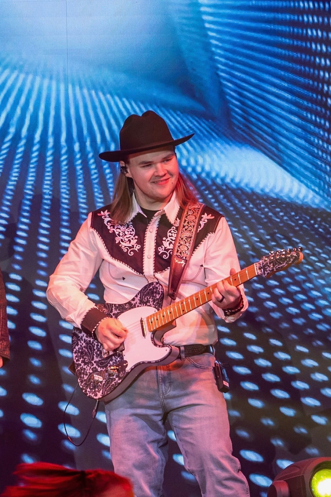 A musician wearing a cowboy hat and western-style shirt plays an electric guitar on a stage with a blue light background.
