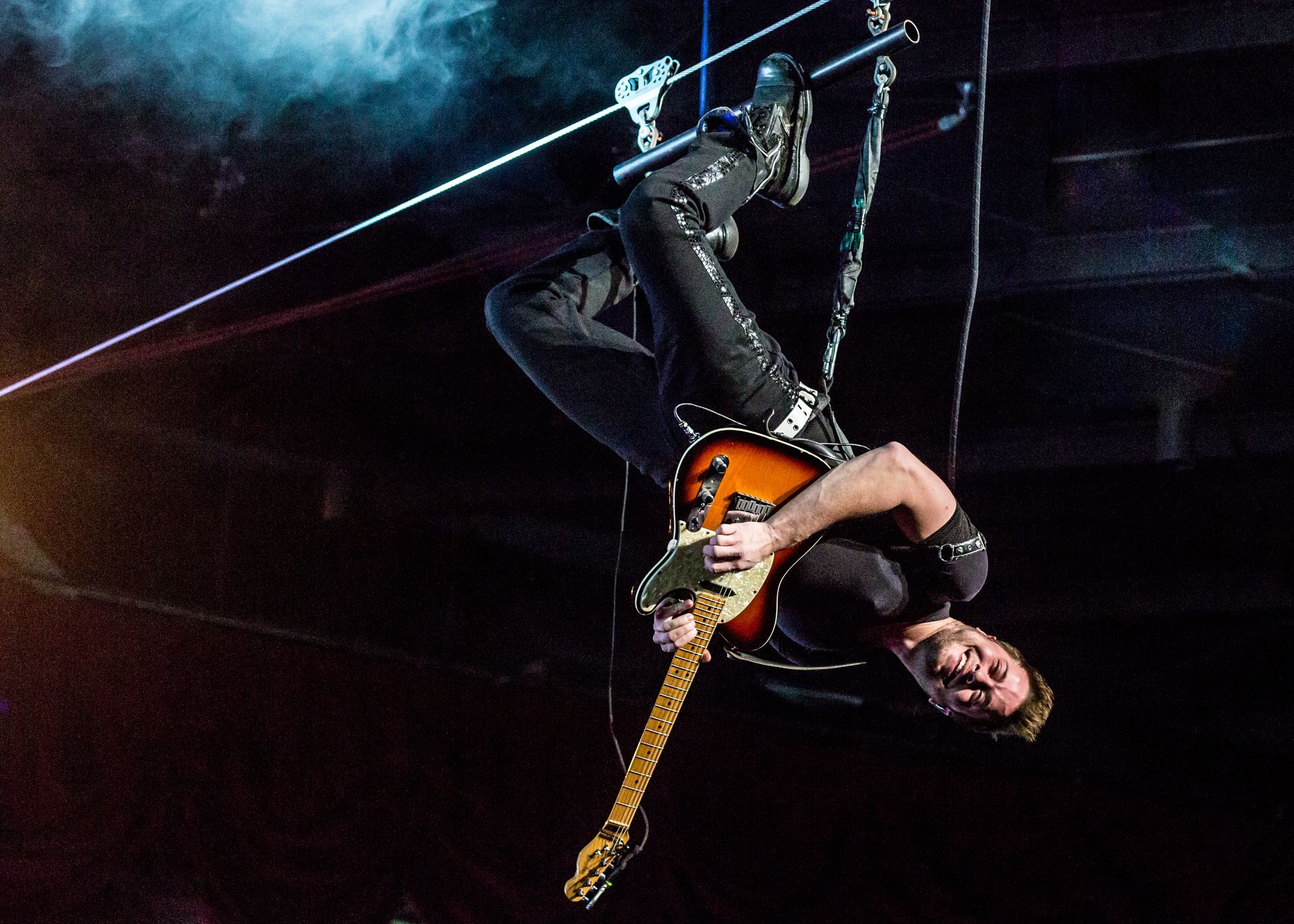 A musician hangs upside down from a zipline while playing an electric guitar during a stage performance.