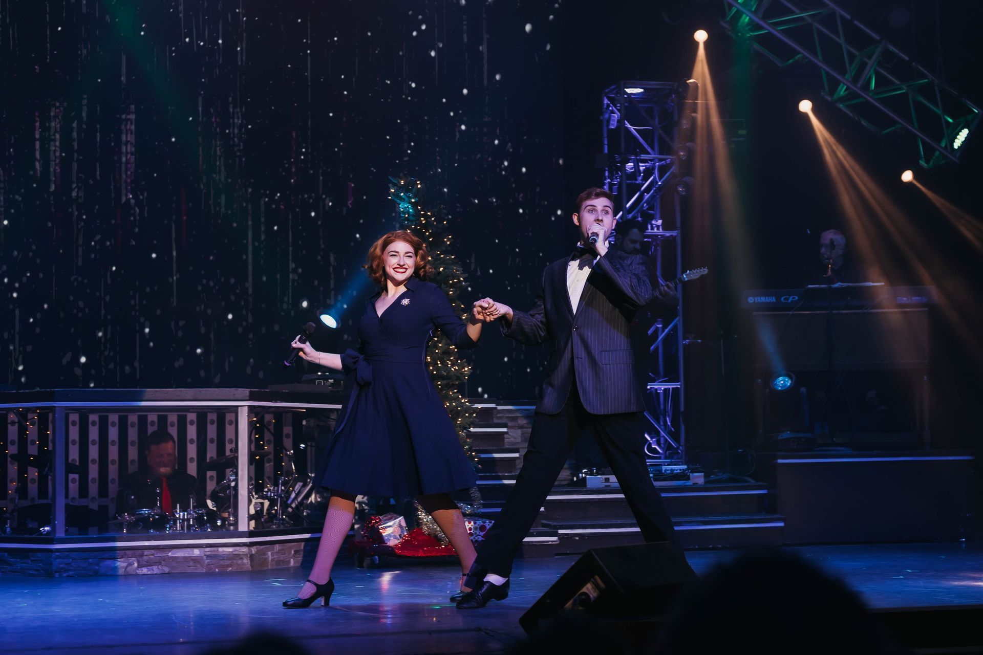 A couple in formal attire performs a dance on a stage with festive lighting and a decorated tree in the background.