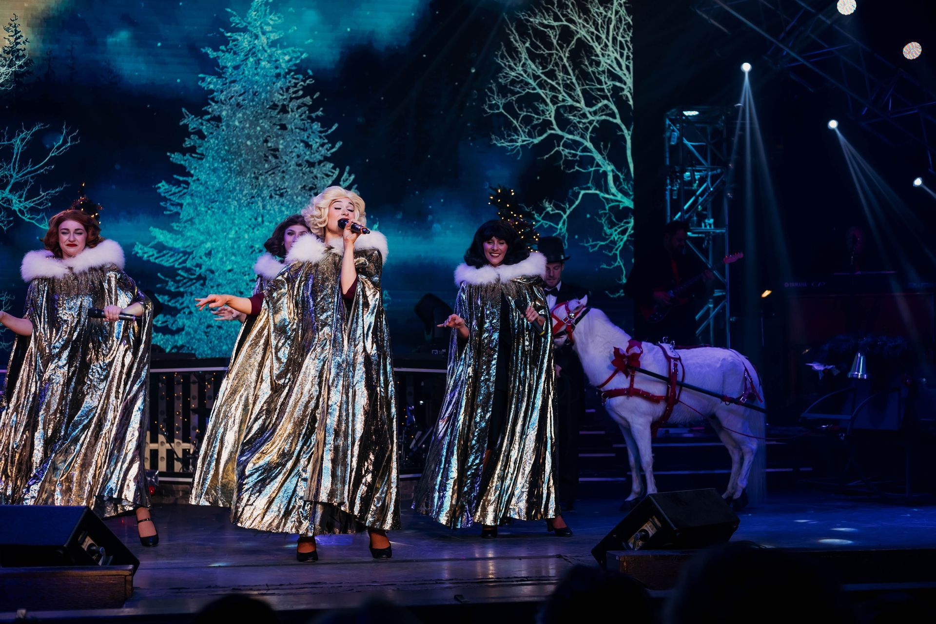 Four performers in shimmering gold capes with white fur trim sing on a stage with a white pony and winter-themed backdrop.