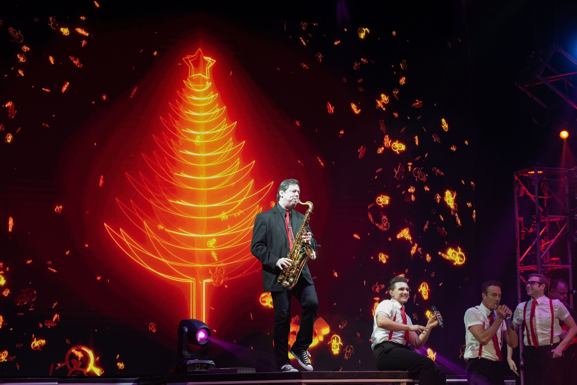 A saxophonist performs on stage in front of a large neon Christmas tree backdrop, with two musicians standing nearby.