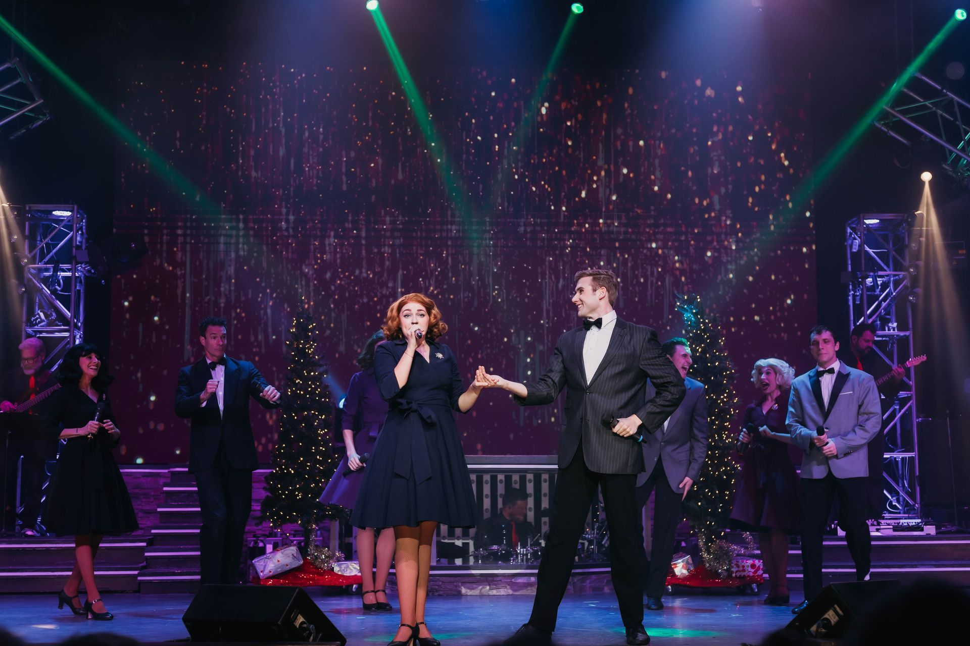 A stage performance featuring a singing duo holding hands center stage, surrounded by a band and festive decor.