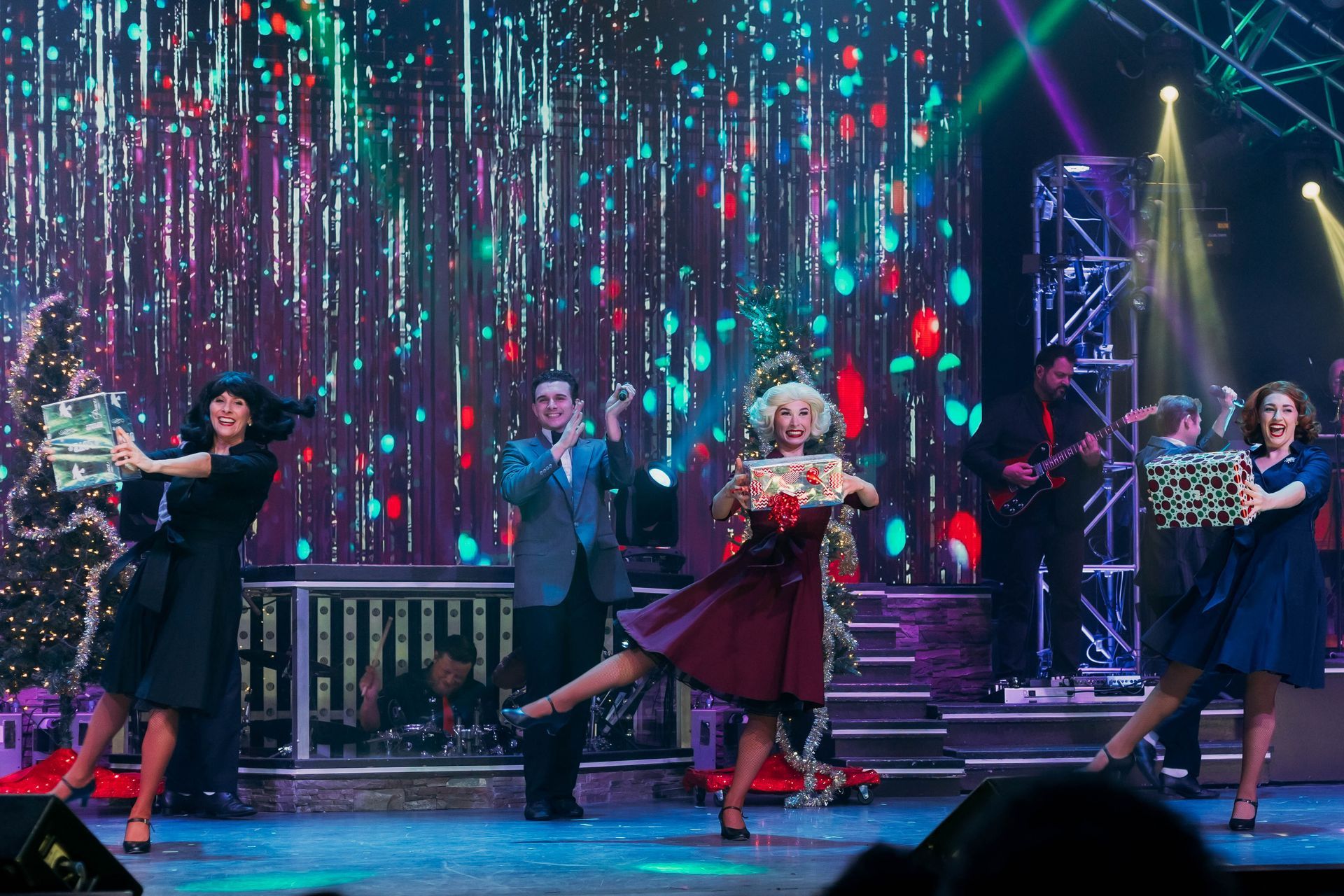 Performers in vintage attire dance on a stage with a sparkling, colorful backdrop, holding wrapped gifts.