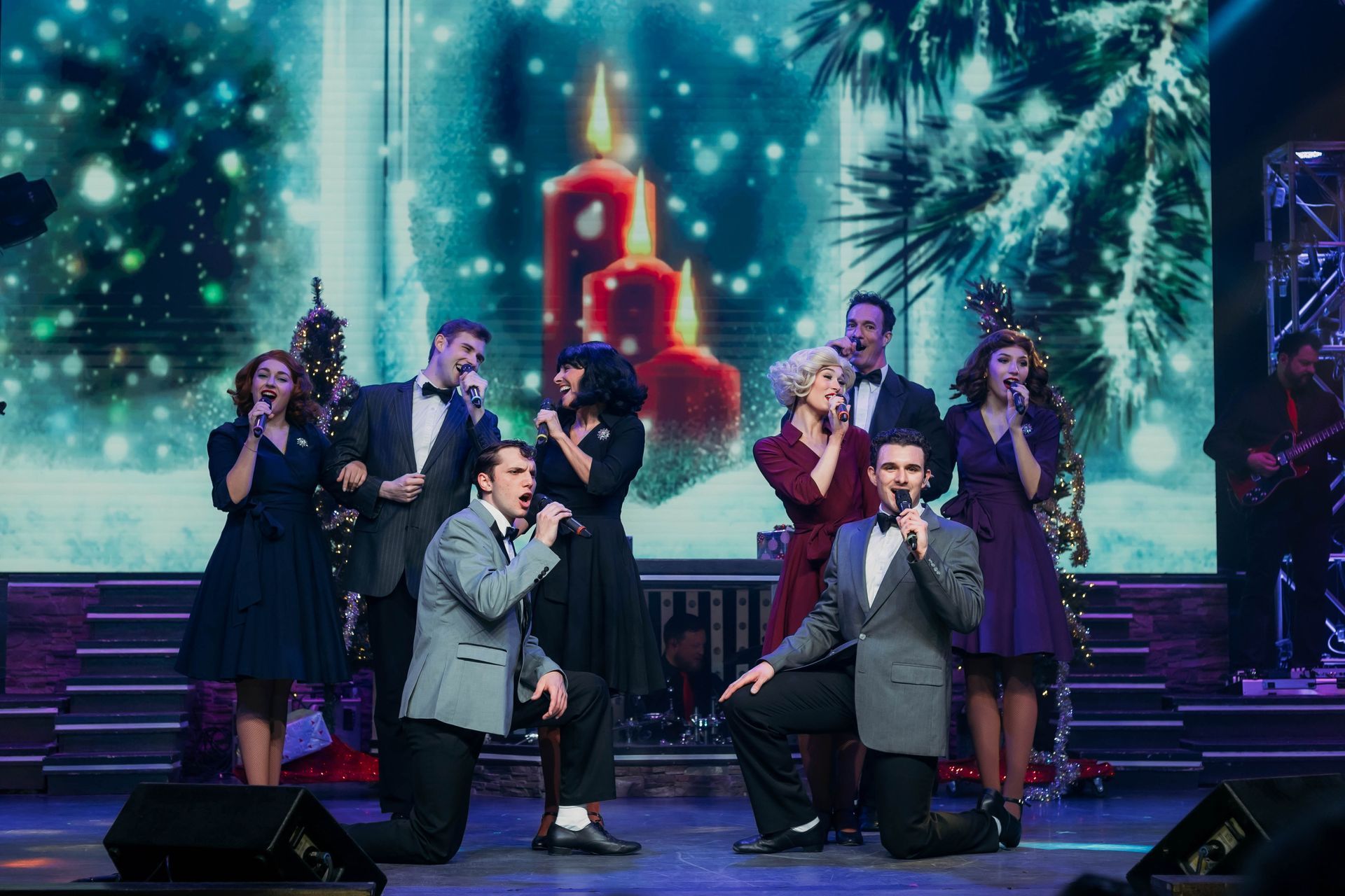 A group of performers in formal attire singing on stage with a backdrop of a snowy forest and lit candles.