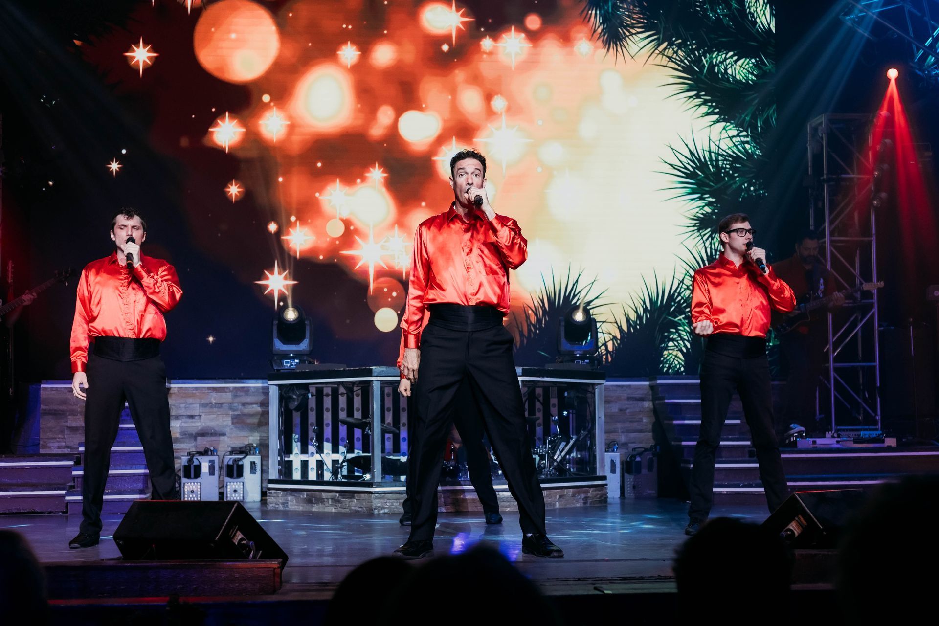 Three singers in matching red shirts and black trousers perform on a stage with a blurred, glowing light background.