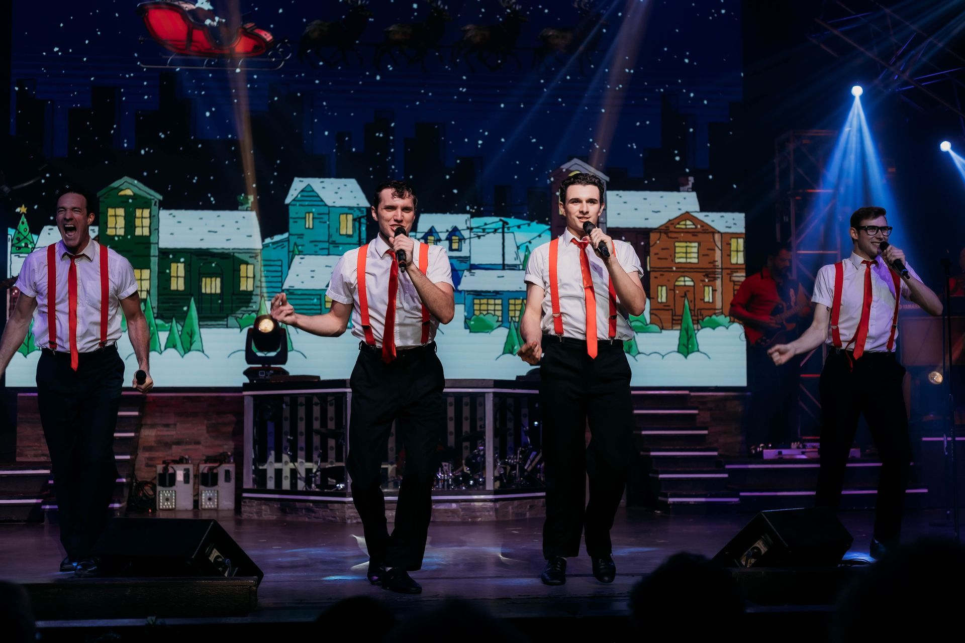 Four singers in white shirts and red suspenders perform on a stage with a festive, winter-themed digital backdrop.