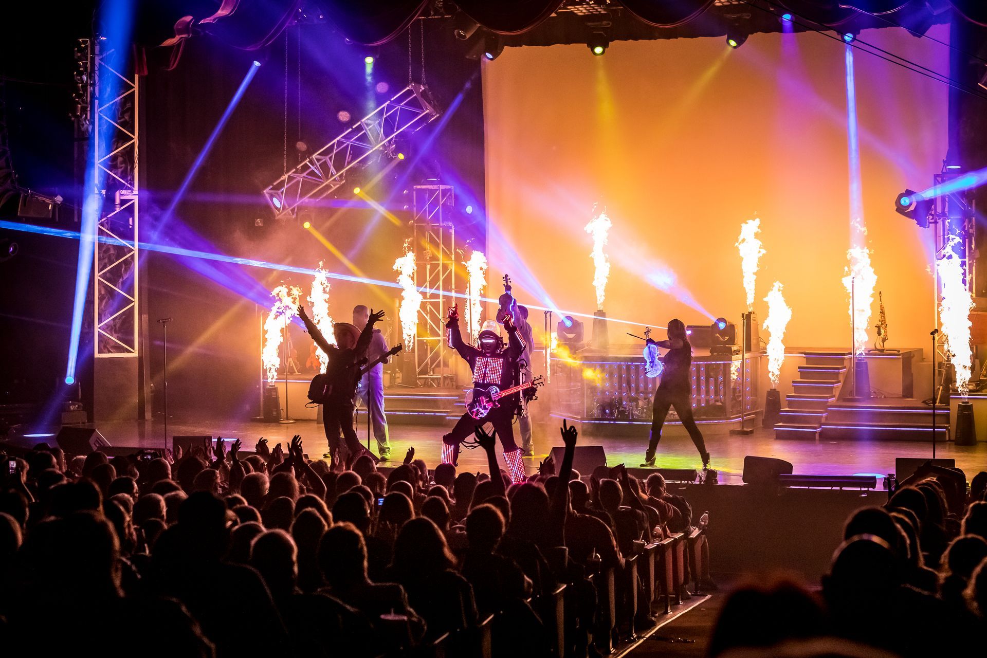 A rock band performs on stage with pyrotechnics, bright blue and yellow spotlights, and a cheering crowd in the foreground.