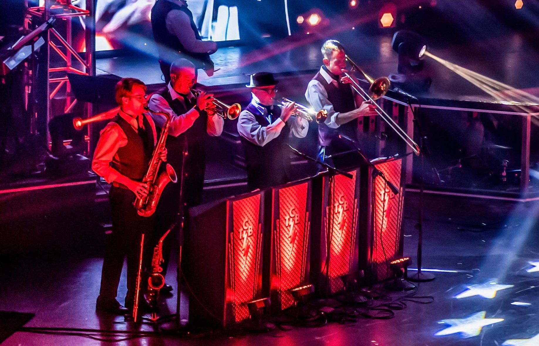 A brass section of four musicians performs on stage, lit by vibrant red light panels and beams from overhead spotlights.
