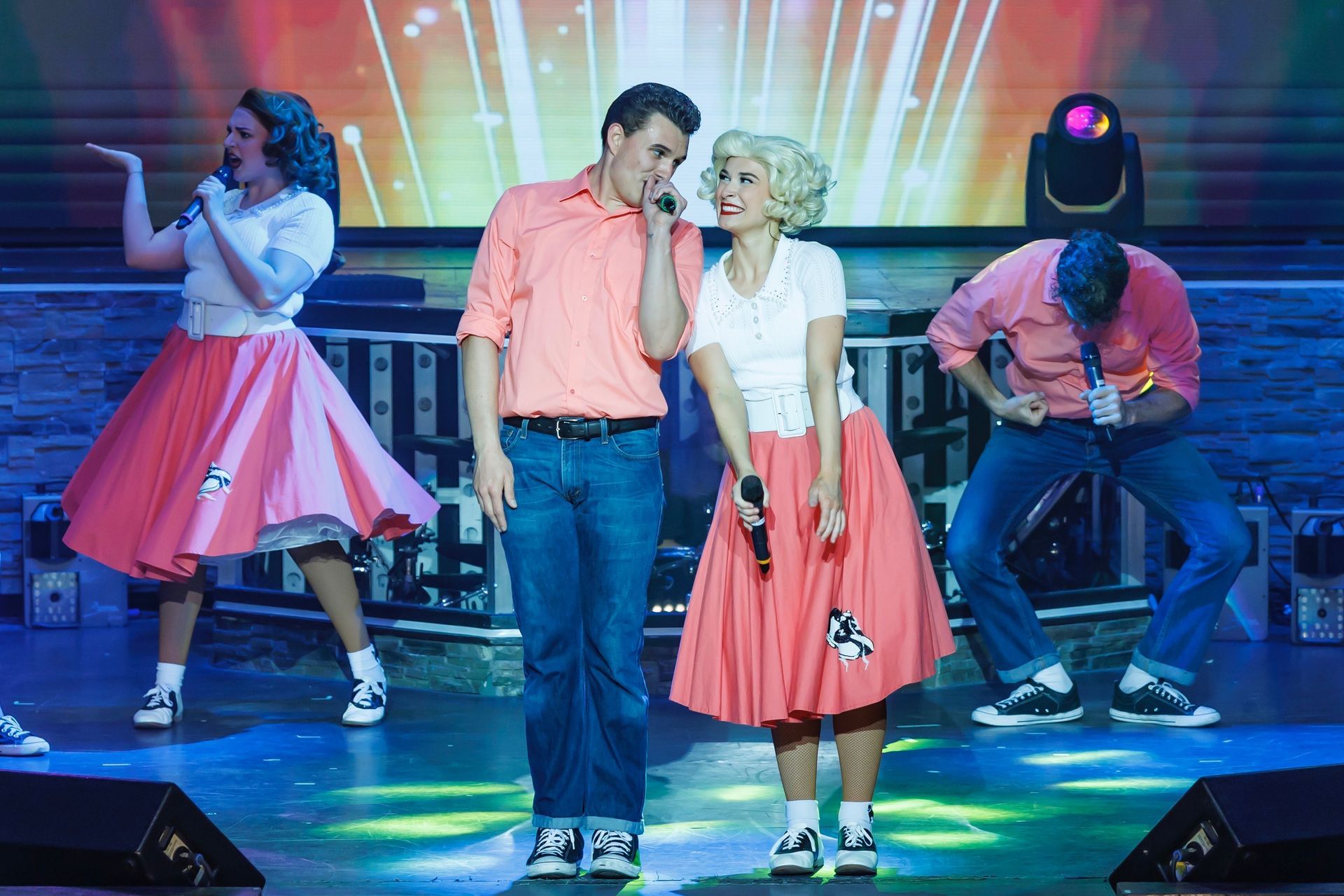 Four performers in 1950s-style clothing sing and dance on a stage with a bright, colorful backdrop.