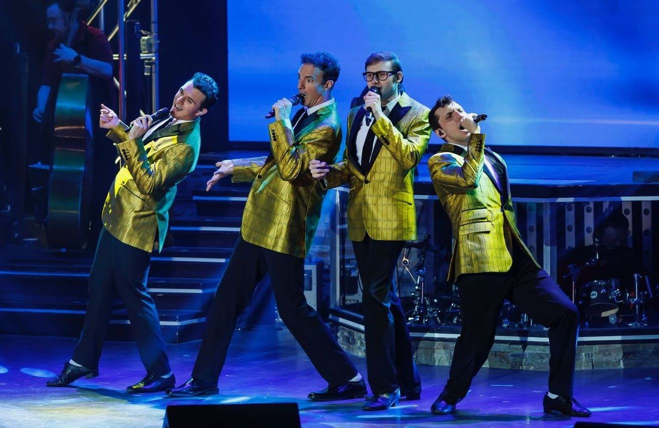 Four performers in matching gold jackets sing into microphones on a stage with blue lighting.