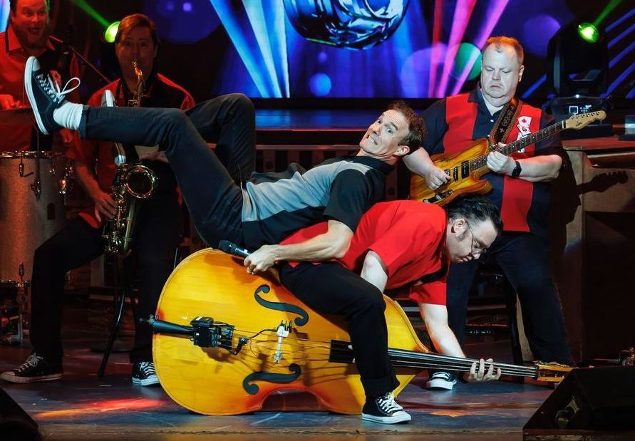 A musician plays an upright bass while a bandmate climbs on top of it, during a lively stage performance.