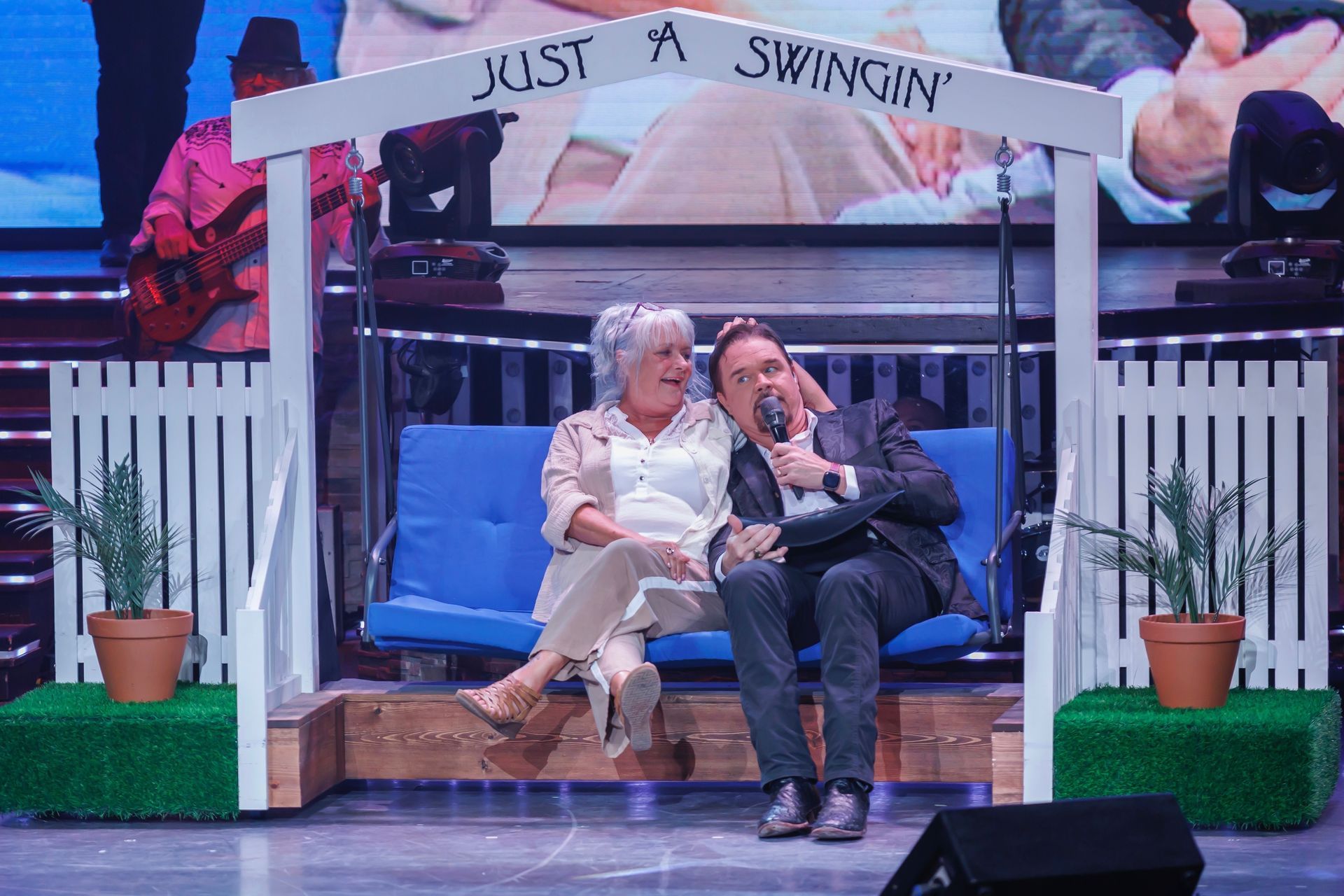 A man and woman sit on a blue porch swing under a white arch labeled 