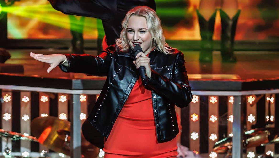 A performer in a red outfit and black leather jacket gestures while holding a microphone on a lit stage.