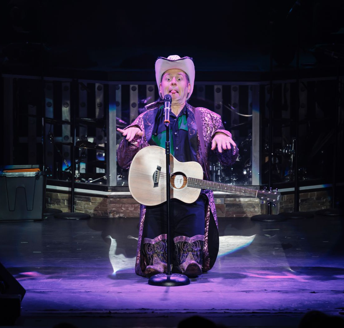 A performer in a purple patterned coat and hat kneels on stage with an acoustic guitar, looking at a microphone.