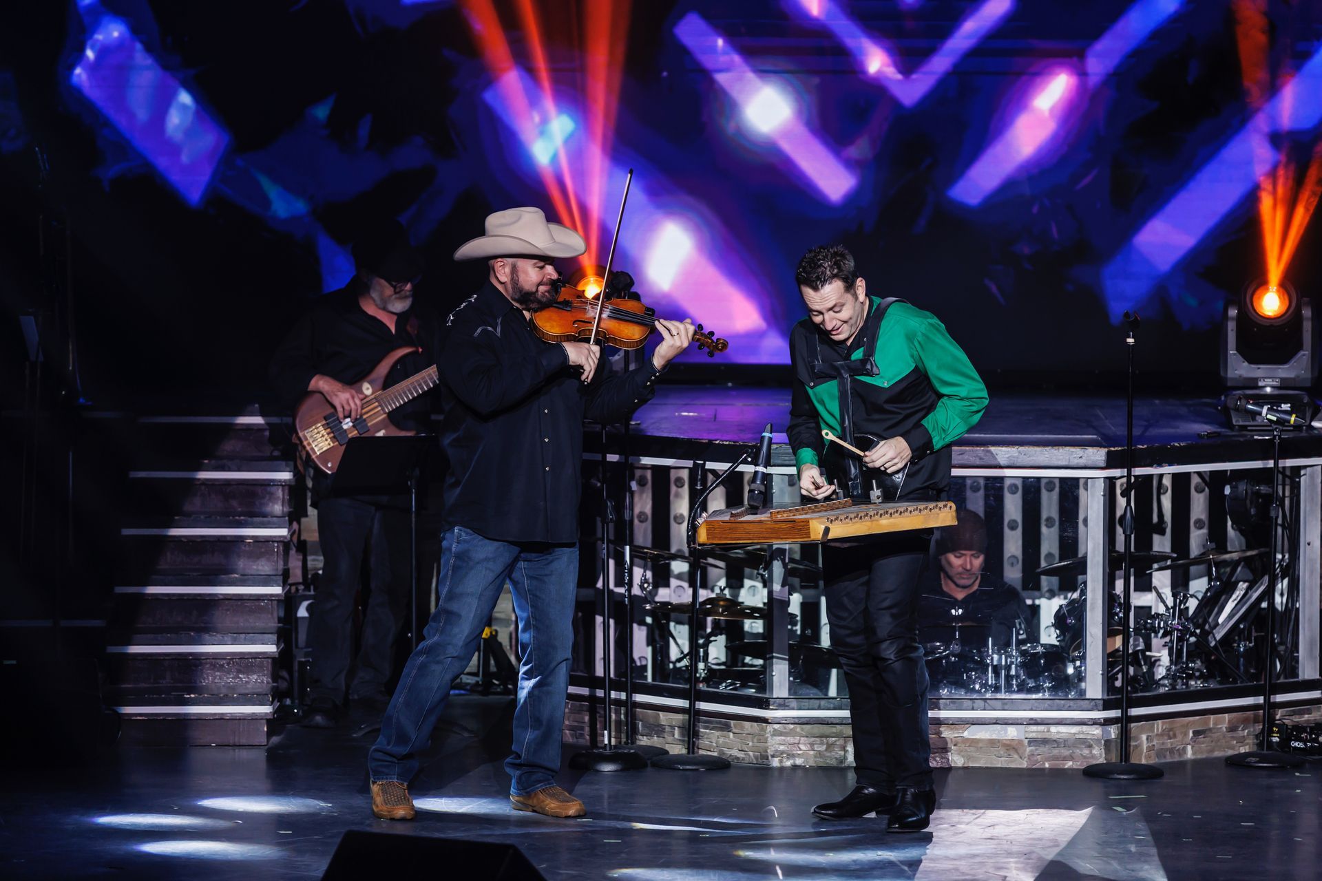 Two musicians perform on stage; one plays a violin while the other plays a hammered dulcimer under bright stage lights.