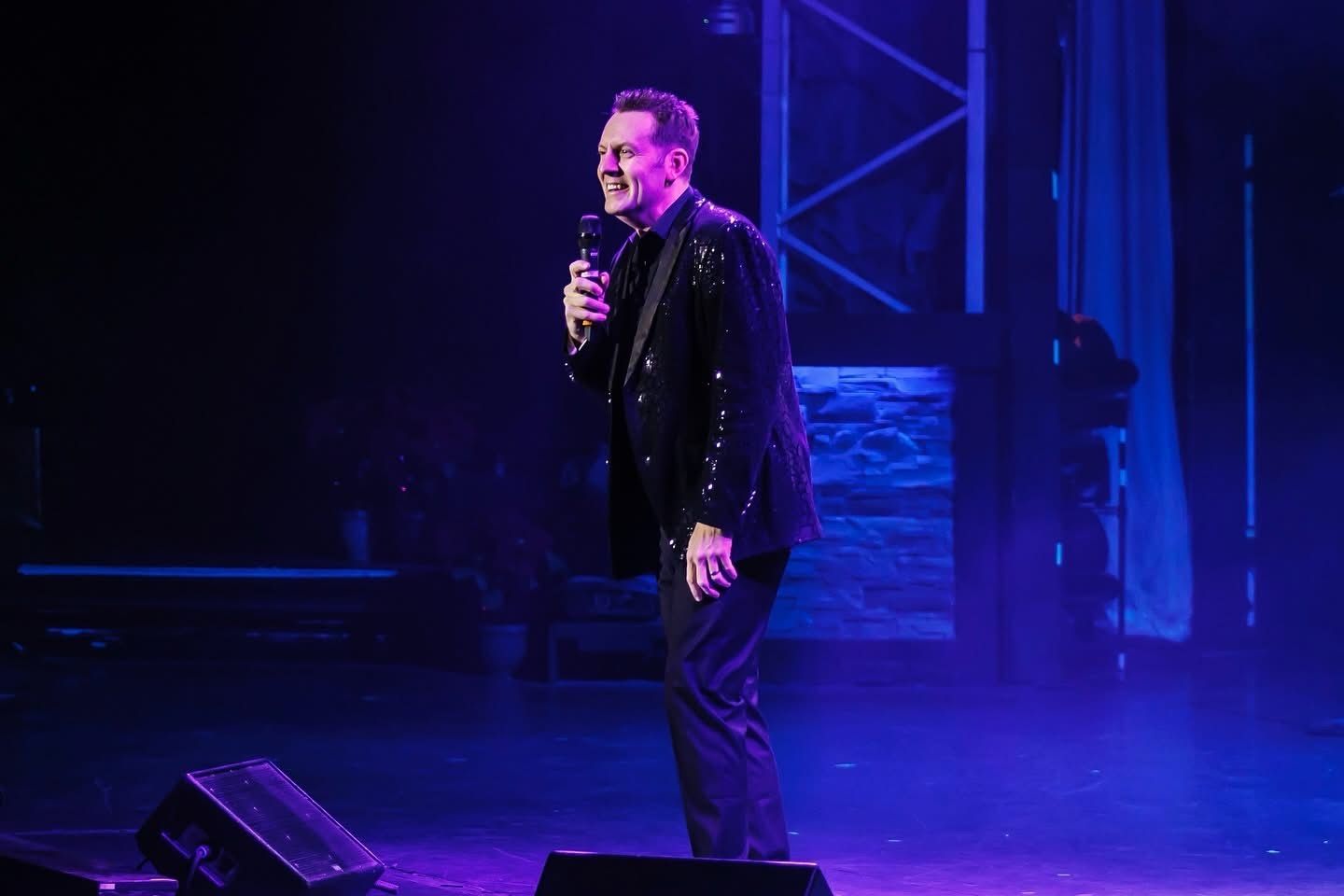 A performer in a sequined jacket holds a microphone on a stage bathed in blue light.