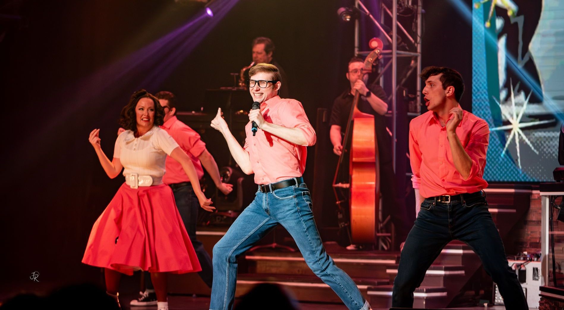A group performing on stage in 1950s-style clothing, with singers and a bass player under bright spotlights.