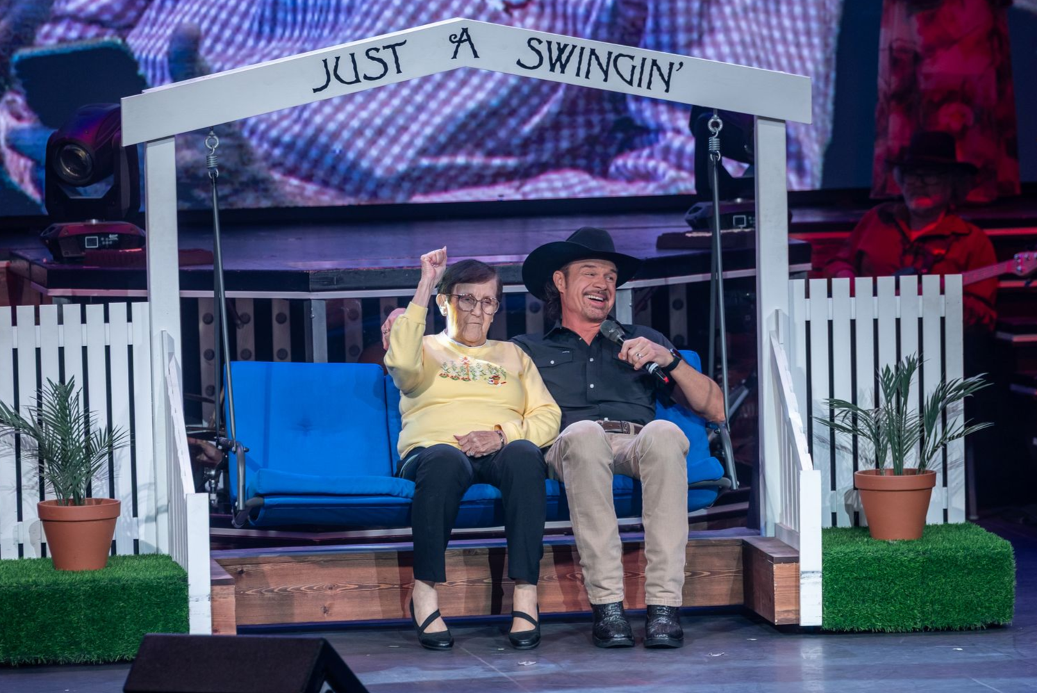 on stage there's a man sitting on a swing in a cowboy hat with an older woman in a yellow sweater who is holding her hand up 