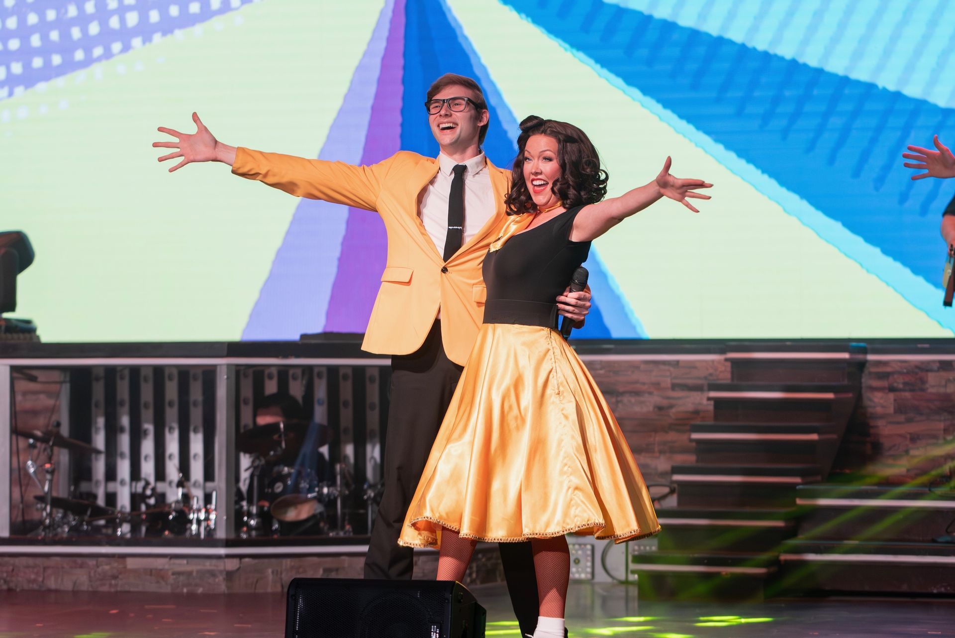 A person in a yellow blazer and a person in a yellow skirt perform on a stage with a bright, colorful background.