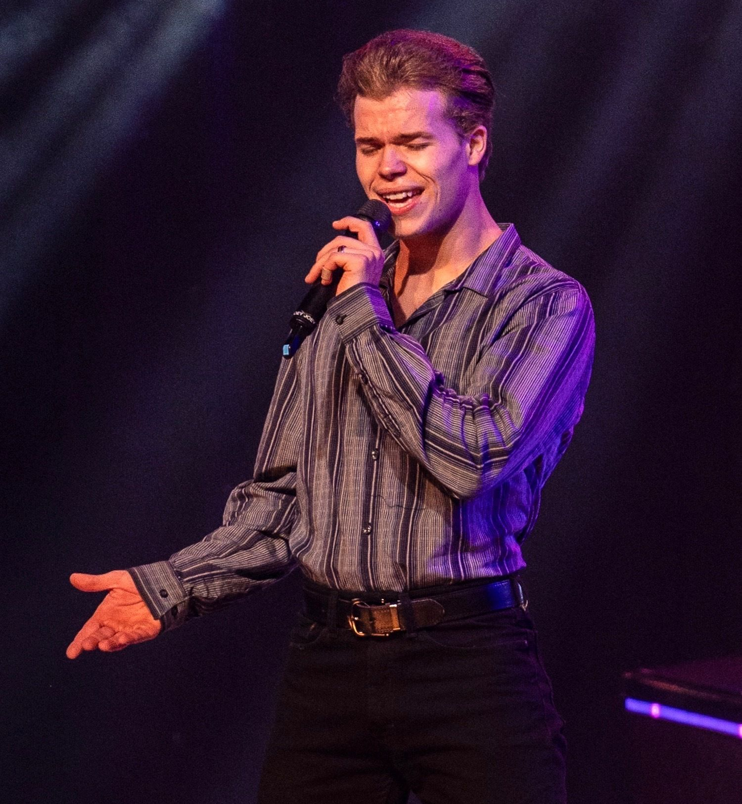 A smiling individual wears a dark green and blue plaid long-sleeved shirt and blue jeans, standing on a stage.