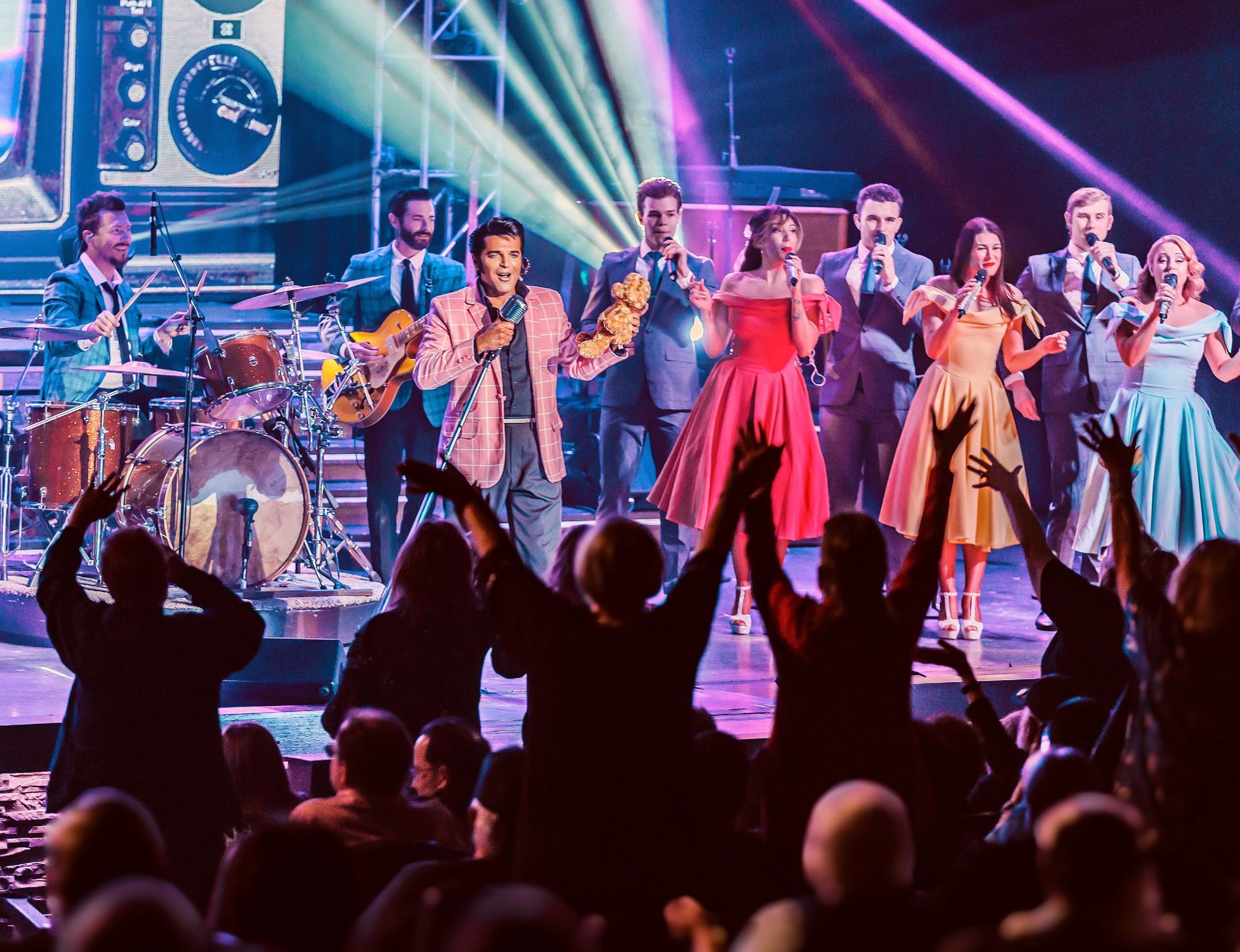 Performers in 1950s-style clothing sing on a stage under bright lights, facing a crowd with arms raised in the foreground.