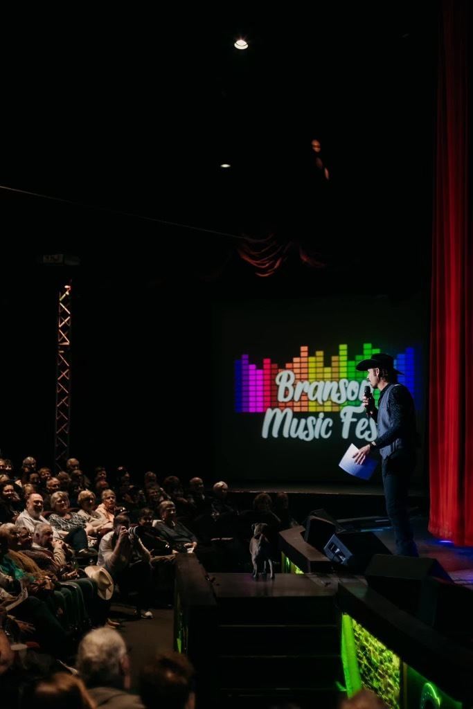 A performer on stage holding a microphone at the Branson Music Fest, with an audience watching from a darkened theater.