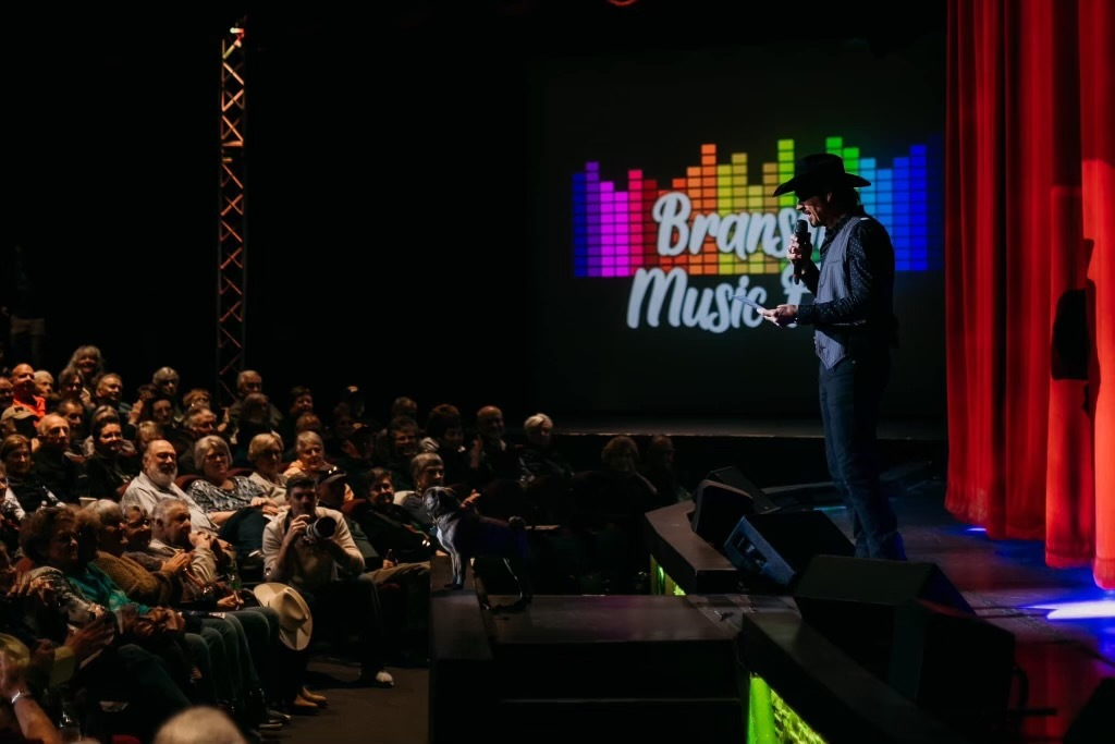 A person in a hat holding a microphone on stage, addressing a large audience in a theater with a colorful logo behind them.