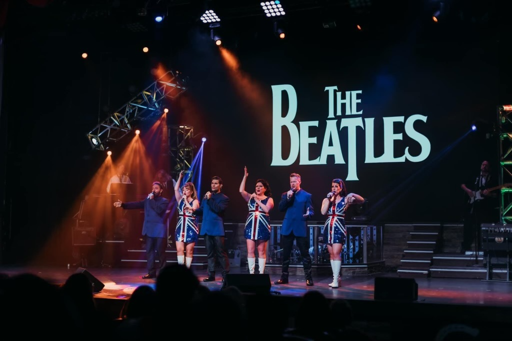 A group of performers singing on a stage in front of a large, glowing "The Beatles" sign.