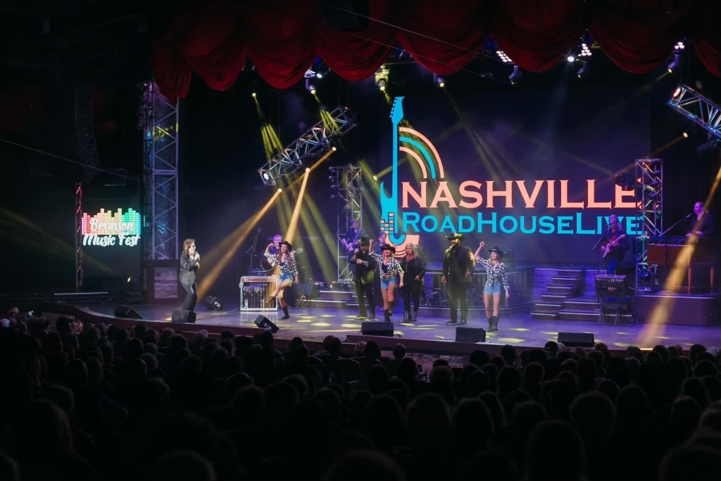 A country music band performs on a stage with neon signs under bright spotlights, viewed from a large, seated audience.