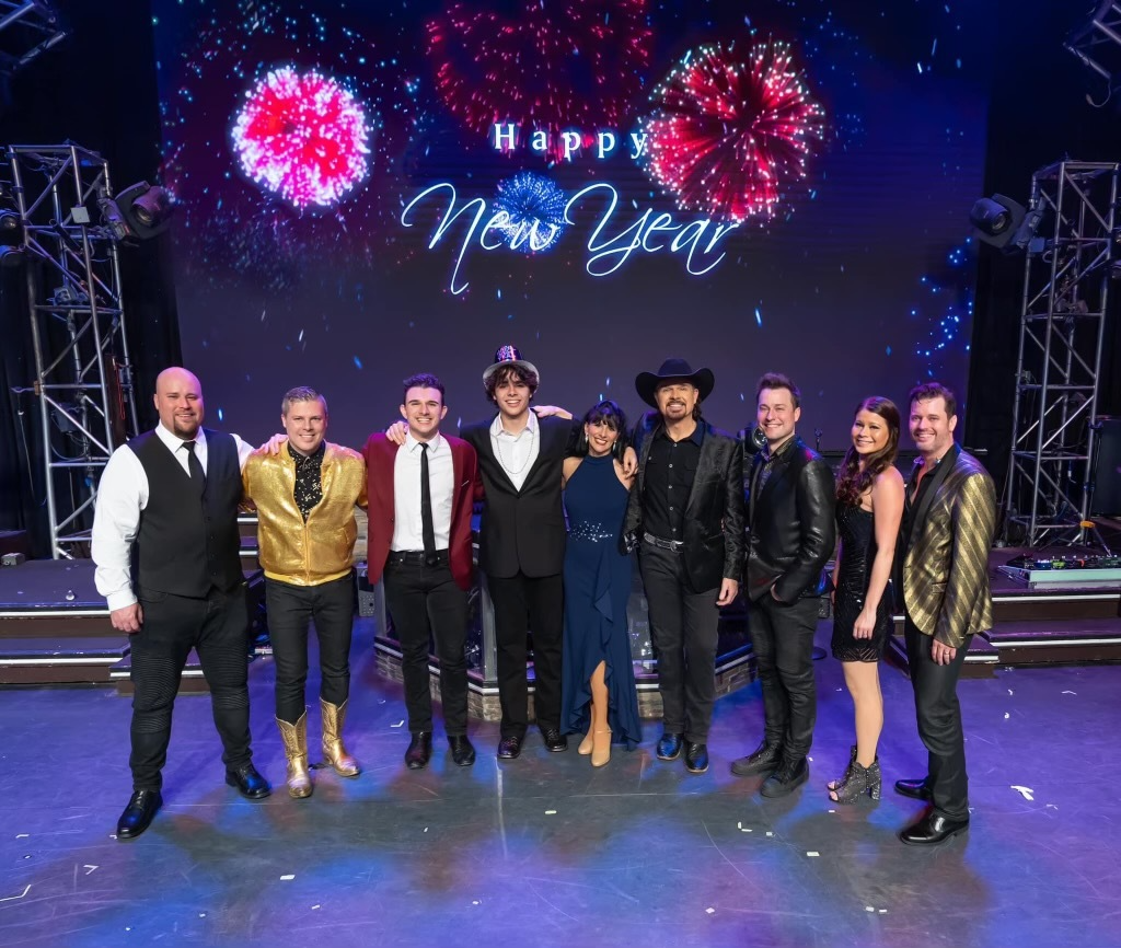 A group of nine people stand on a stage in front of a digital screen displaying "Happy New Year" and fireworks.