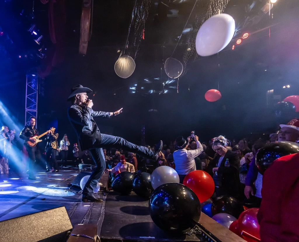 A musician in a cowboy hat performs on stage while lifting a leg, surrounded by balloons over a crowd at a concert.