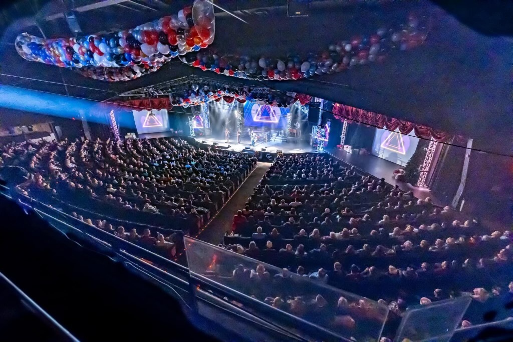 High-angle view of a large, packed auditorium with festive balloons and stage lighting during a performance.