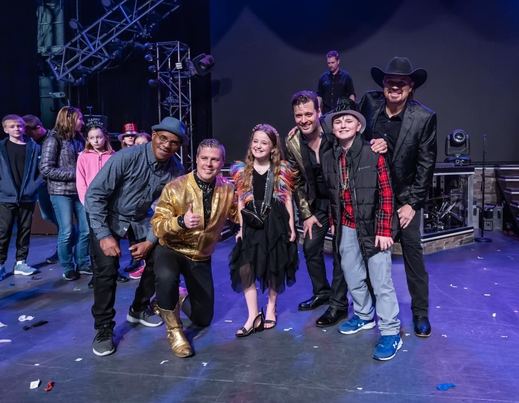 A group of performers in costumes poses together on a stage with a dark backdrop and stage lighting.