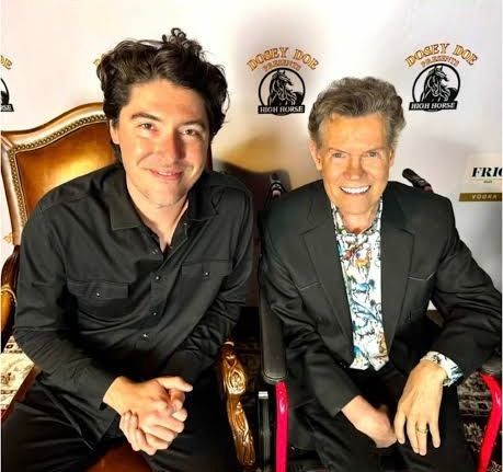 two men sitting next to each other in black shirts/jackets and smiling for a photo with their hands in their laps 