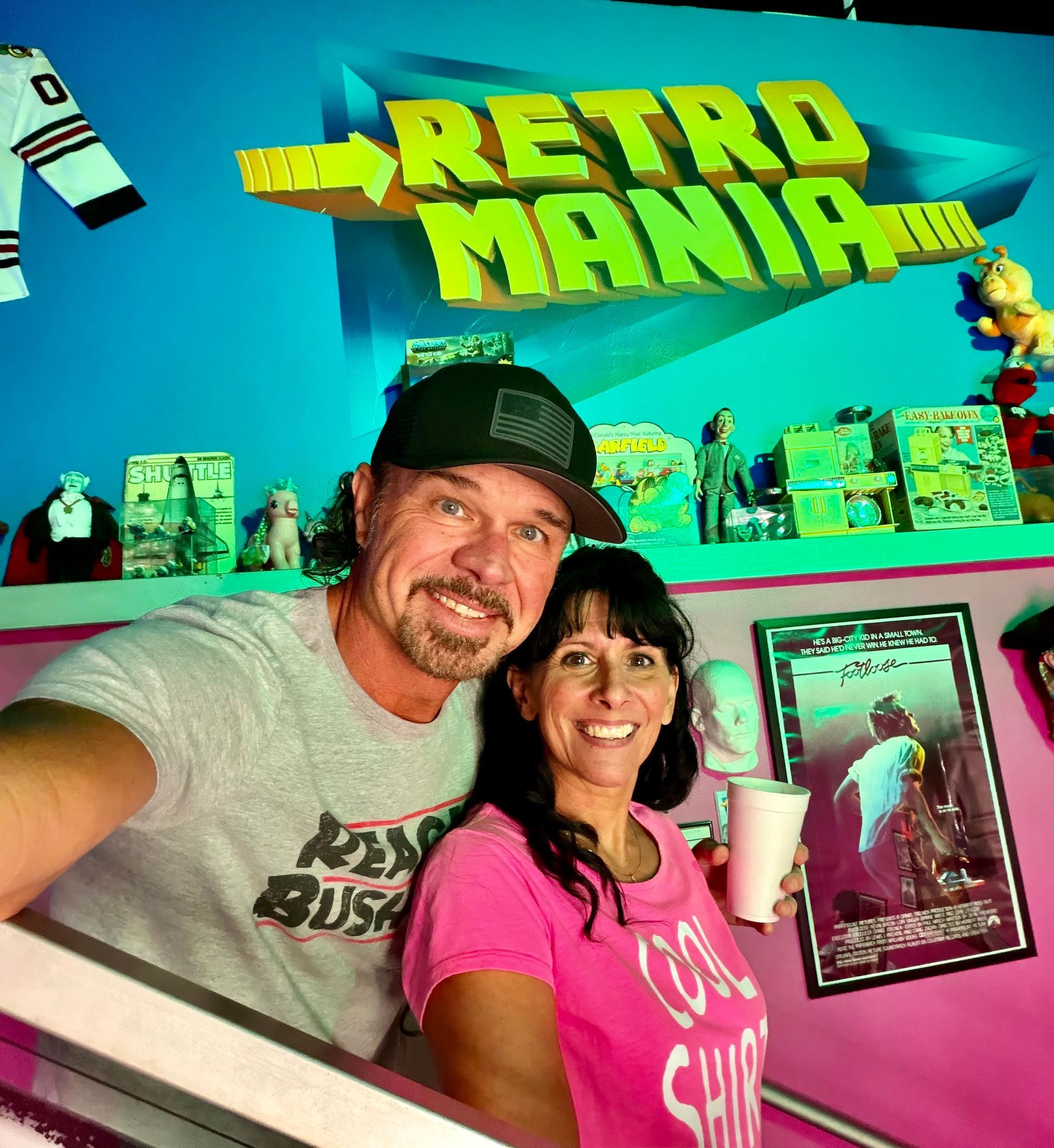 man and woman taking a selfie in front of a wall that says Retro Mania and has old 80's toys in the background 