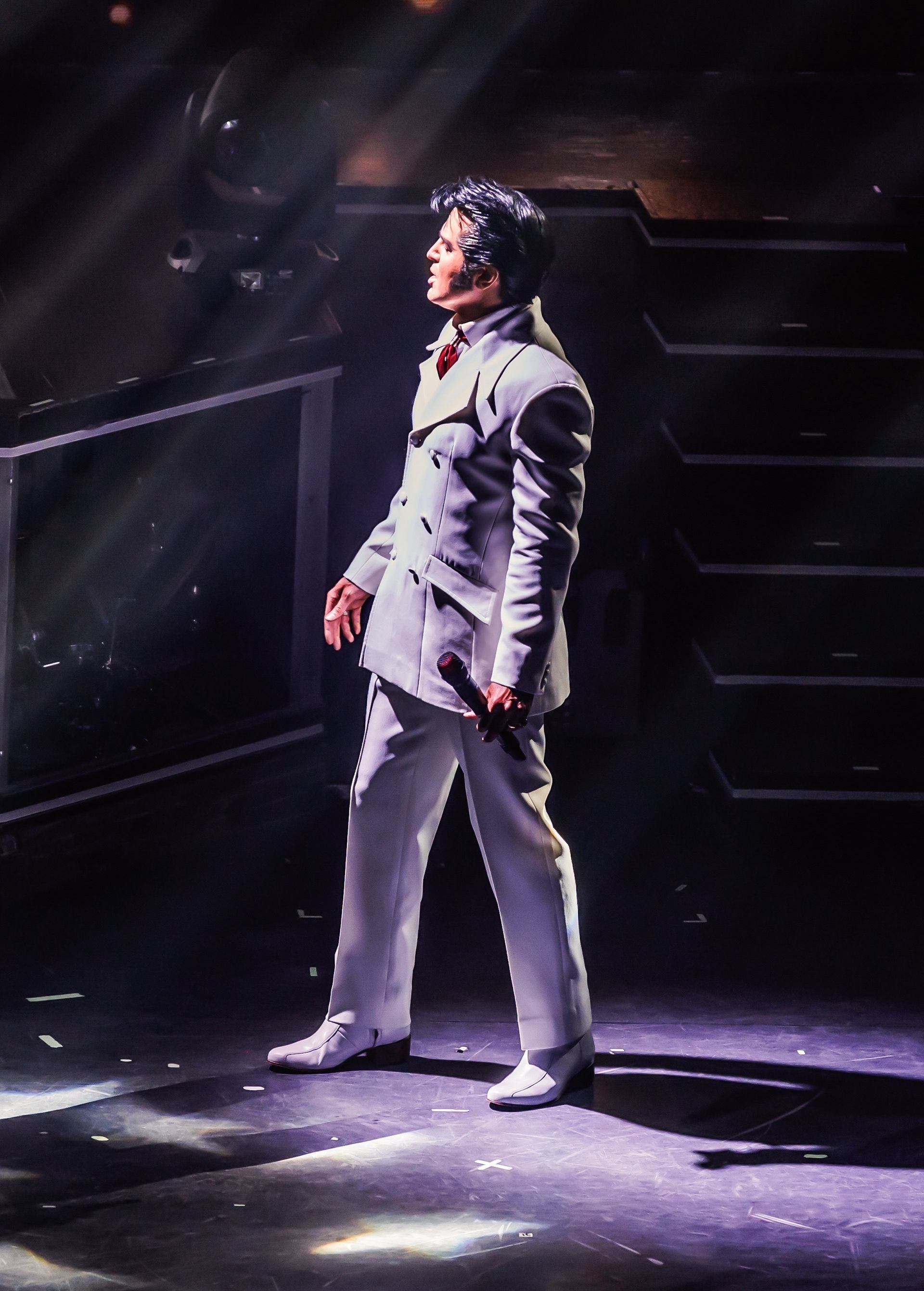 An Elvis impersonator in a white suit and dark pompadour hairstyle stands on a dark stage, illuminated by a spotlight.