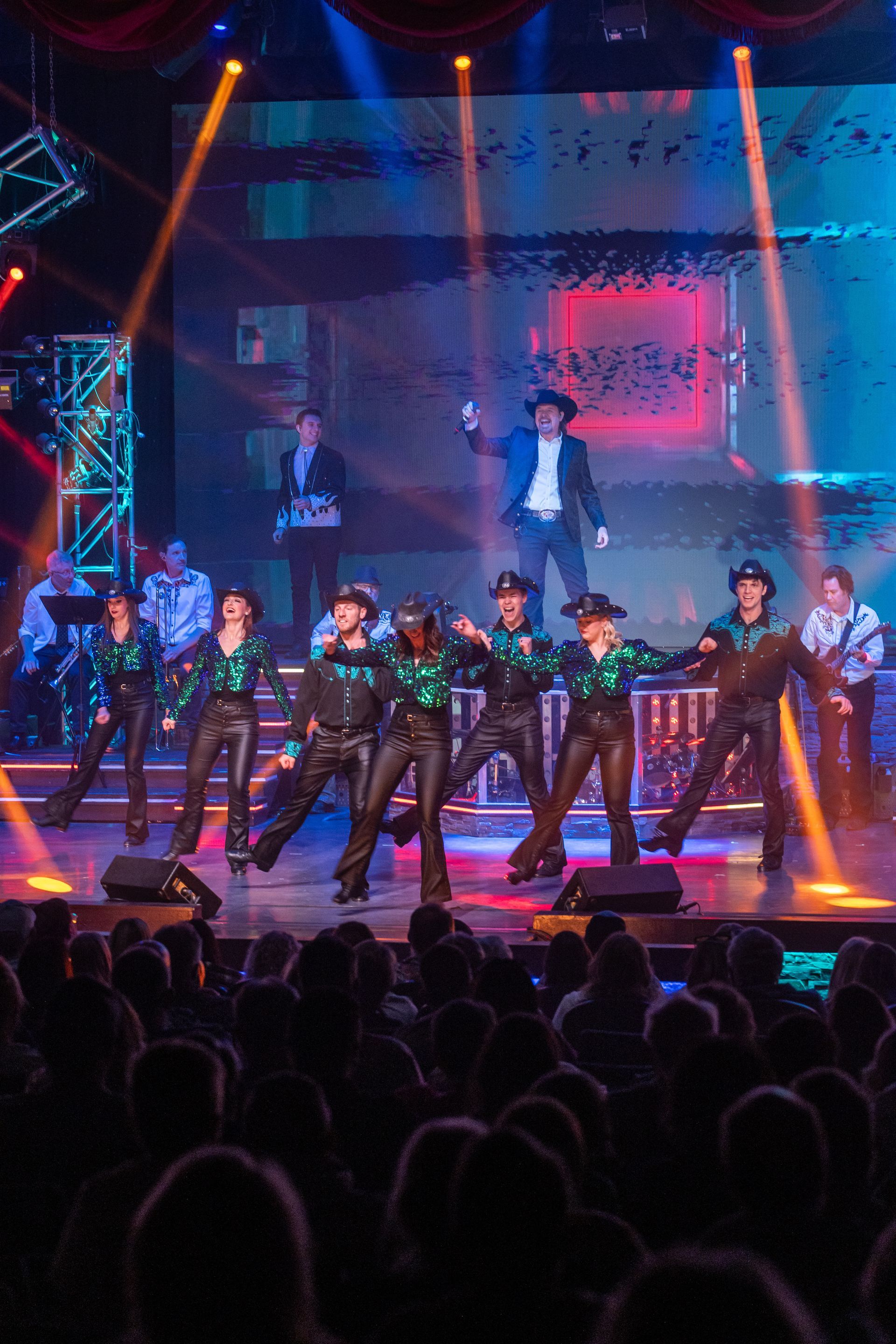 A group of performers in green shirts and black pants dance on a stage under bright spotlights before an audience.