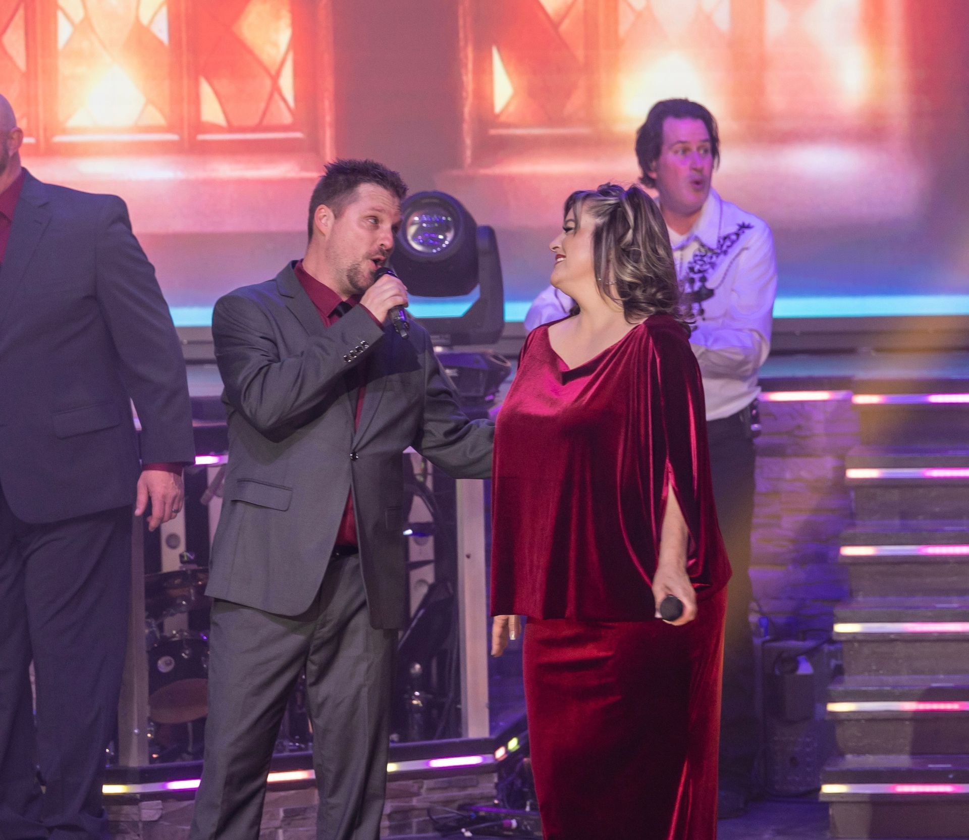 A man and a woman in formal wear singing into microphones on a brightly lit stage with a band in the background.