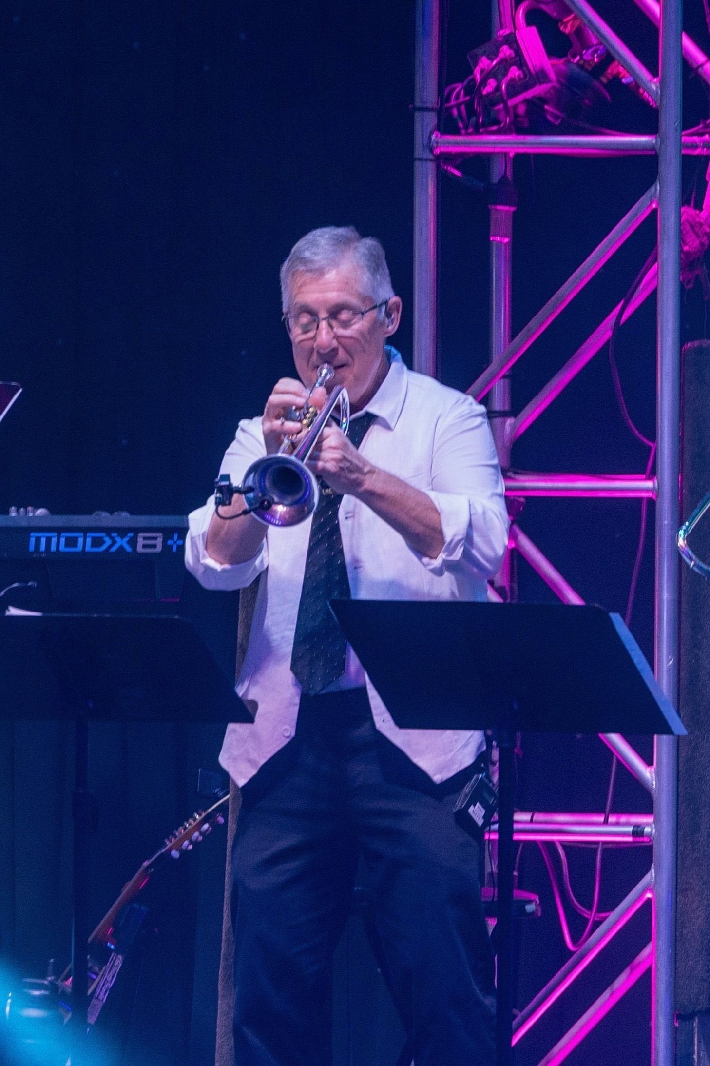 A musician in a white shirt and tie plays a trumpet on stage with purple lighting and metal truss structures.
