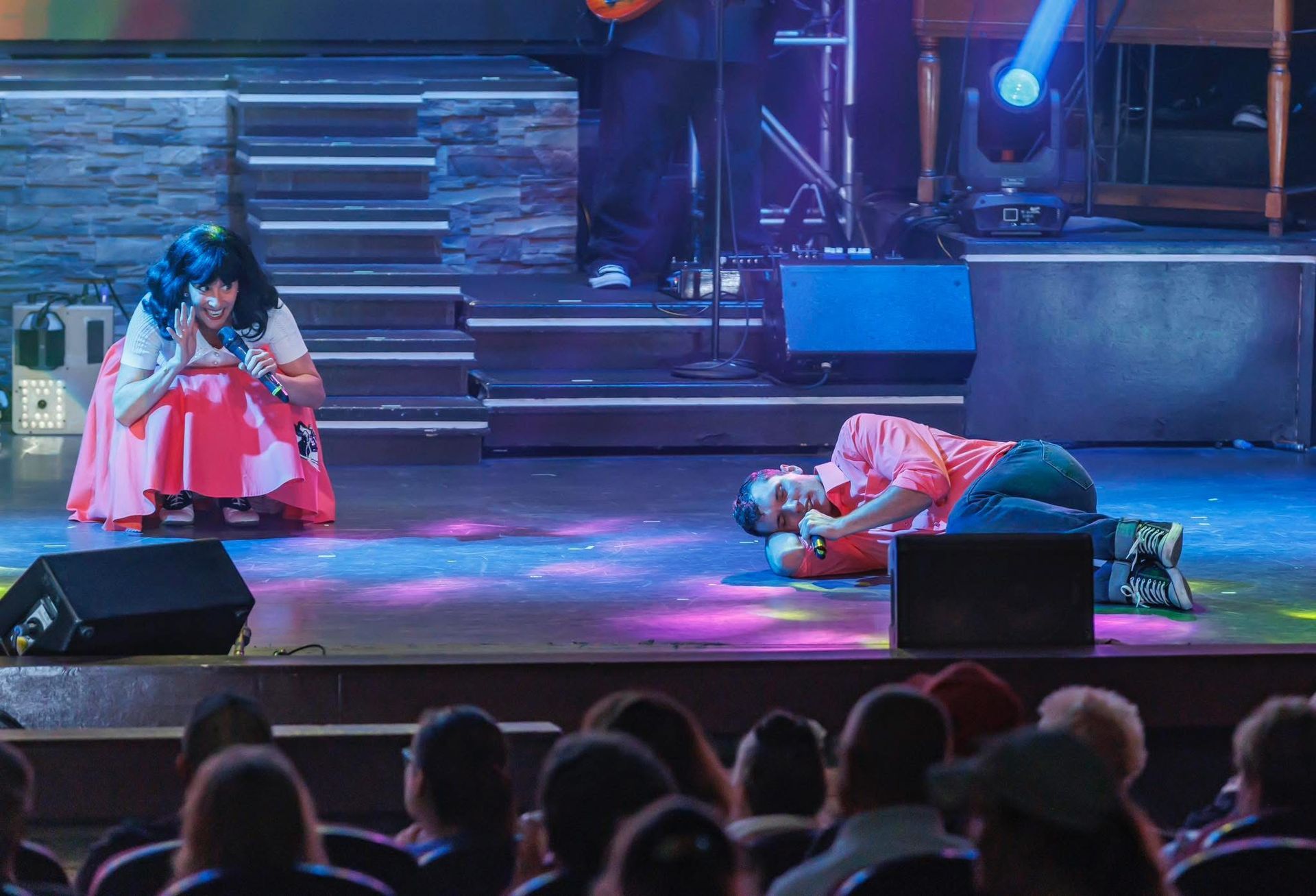 Two performers on a stage: one kneeling in a pink skirt, the other lying on the floor, both holding microphones.
