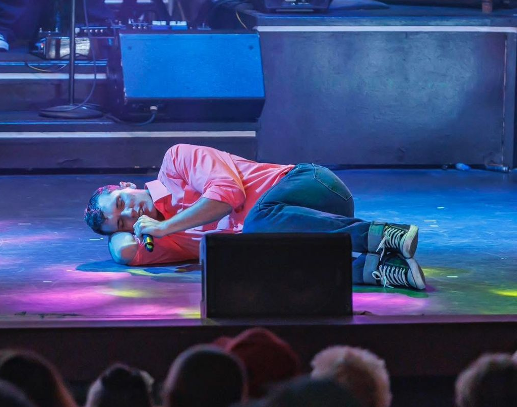 Two performers on a stage: one kneeling in a pink skirt and one lying on the floor, both holding microphones.
