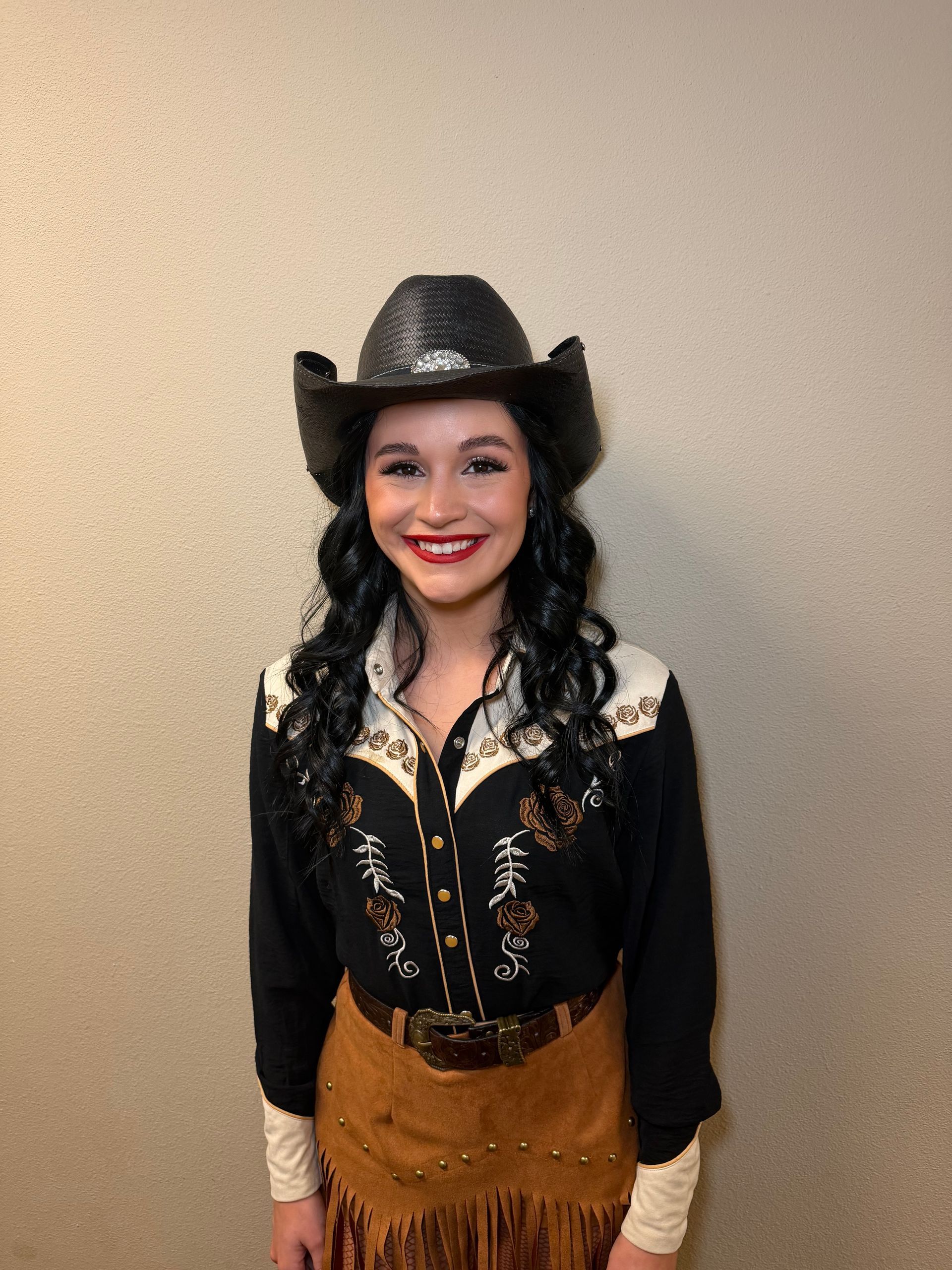 A person wearing a black cowboy hat, a black embroidered western shirt, and a brown suede fringe skirt smiles.