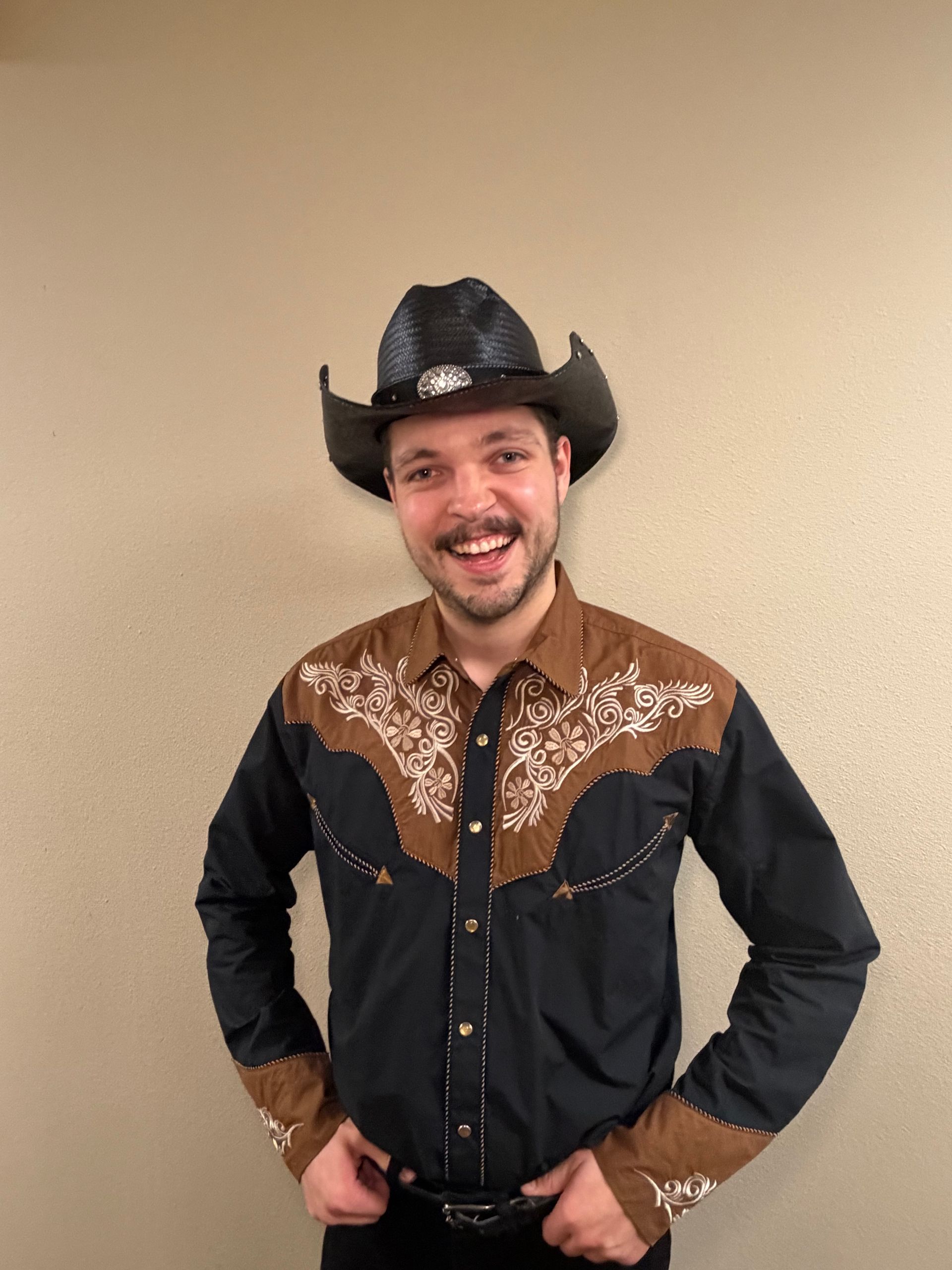 A smiling person wearing a black cowboy hat and a brown and black Western-style shirt with embroidery, standing indoors.