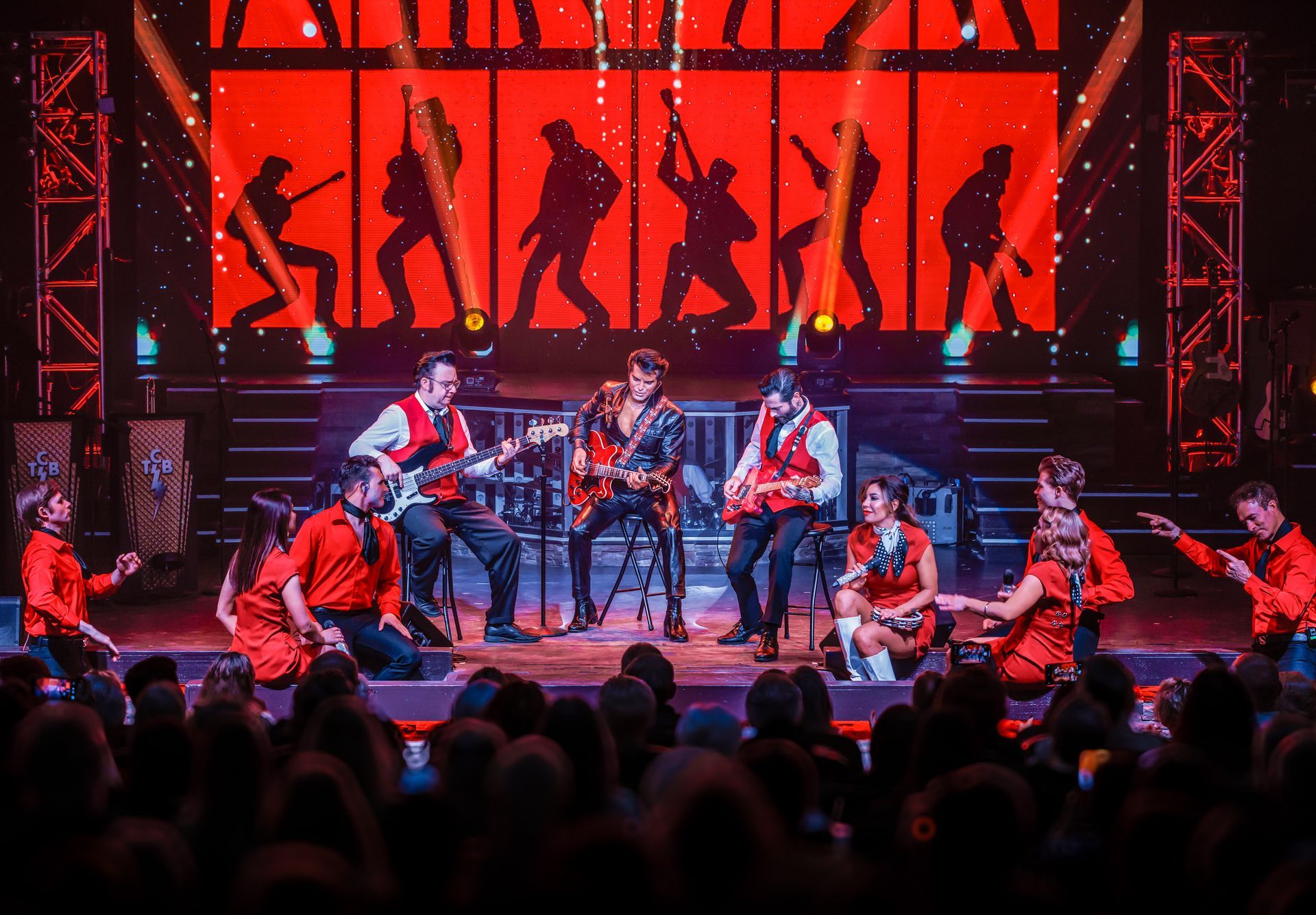 A band performs on a stage with a bright red backdrop, musicians playing instruments, and performers in red outfits.